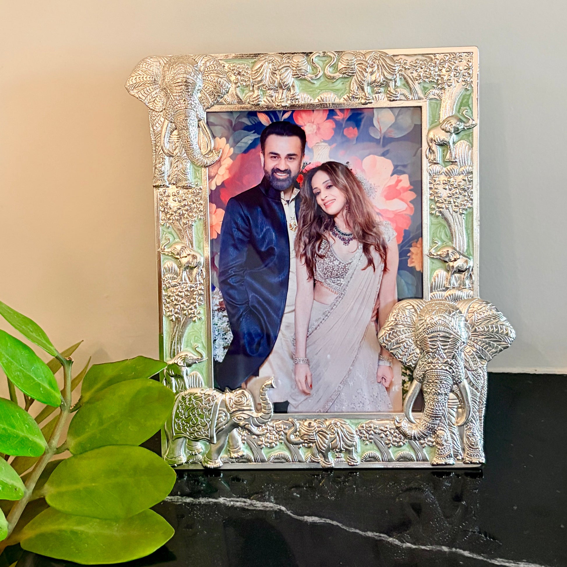 An ornate silver-toned picture frame with elephant and floral carvings, holding a printed photo of a couple, displayed on a black marble surface next to green leaves against a beige wall.