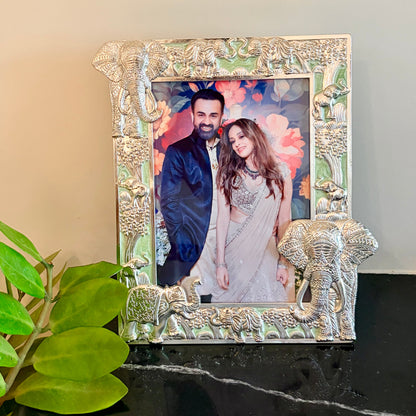 An ornate silver-toned picture frame with elephant and floral carvings, holding a printed photo of a couple, displayed on a black marble surface next to green leaves against a beige wall.